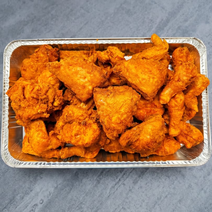 Golden-brown fried chicken pieces fill a rectangular aluminum foil tray on a gray surface.