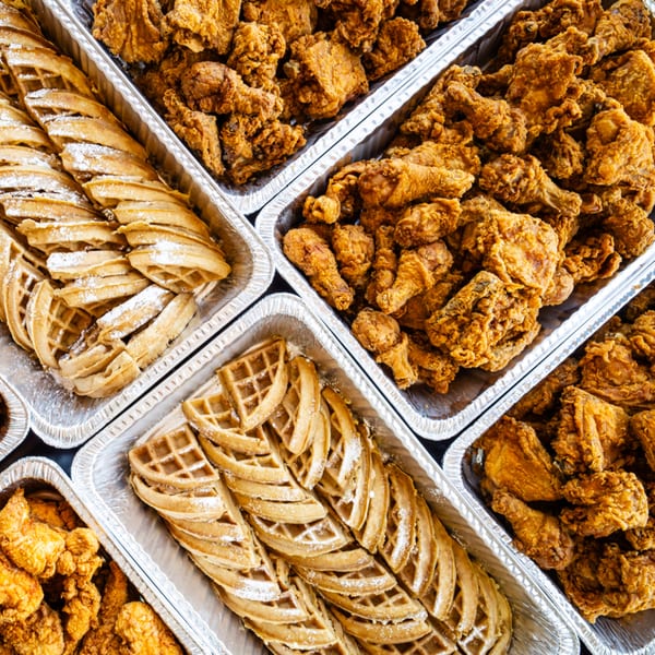 Multiple foil trays filled with golden-brown fried chicken and powdered sugar-dusted waffles.