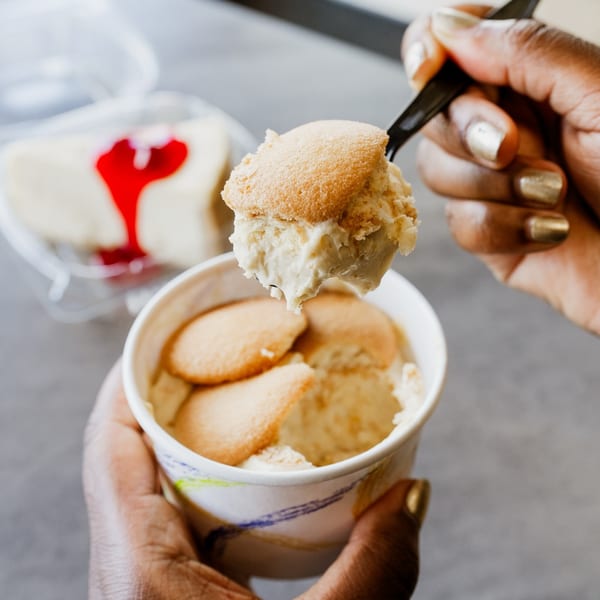 Hands holding a cup of banana pudding with vanilla wafers, cheesecake in background.