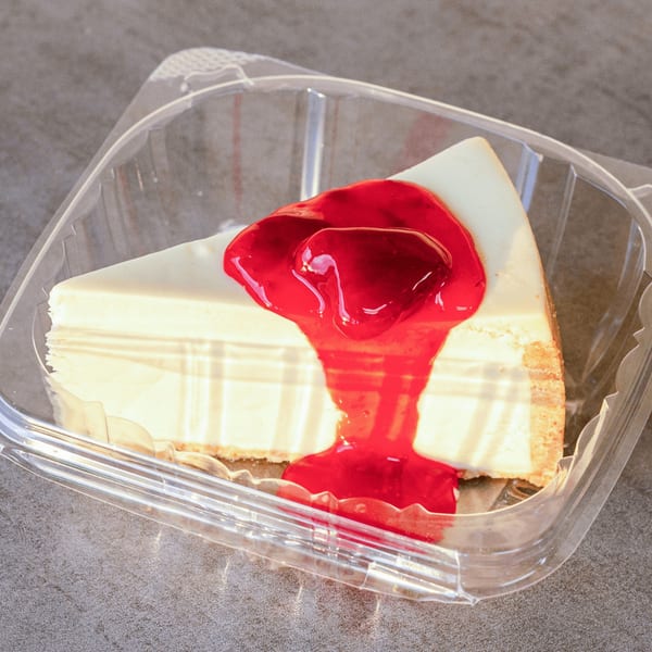 A slice of cheesecake with strawberry topping in a clear plastic to-go container.