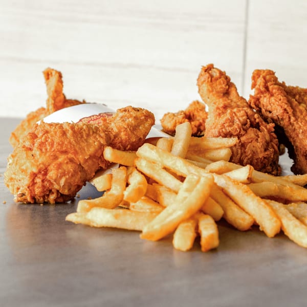 Crispy fried chicken pieces and golden french fries on a gray surface, with a white sauce cup.