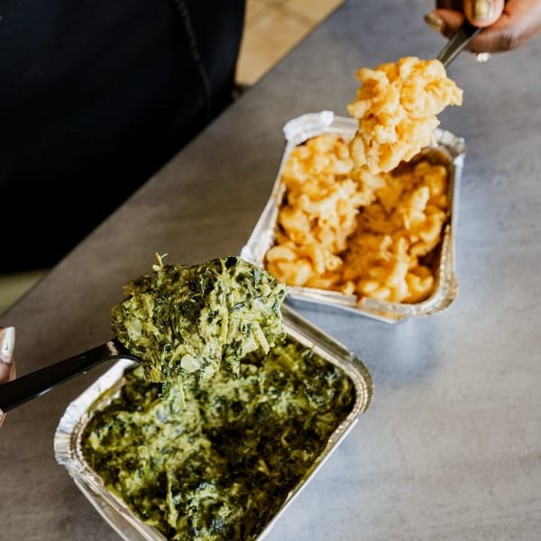 Hands scooping creamy mac and cheese and green collard greens from foil containers.